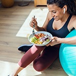 Woman wearing workout clothes leaning on medicine ball eating salad